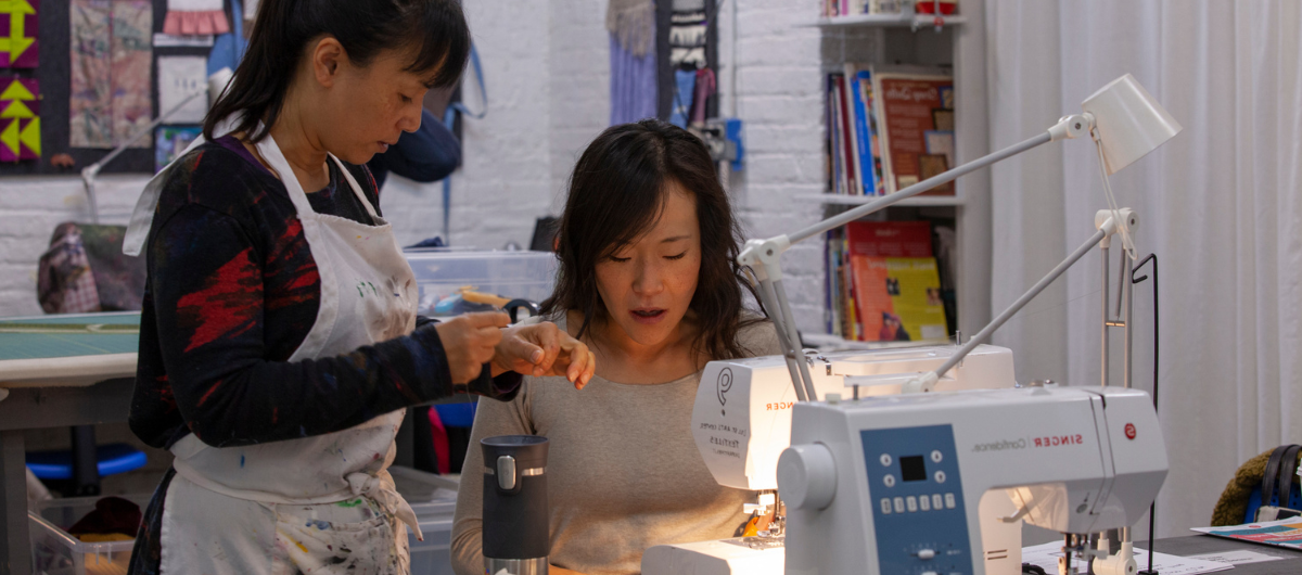 A teacher in a long sleeve black sweater and white apron, standing, assists a student with long hair, sitting at a sewing machine