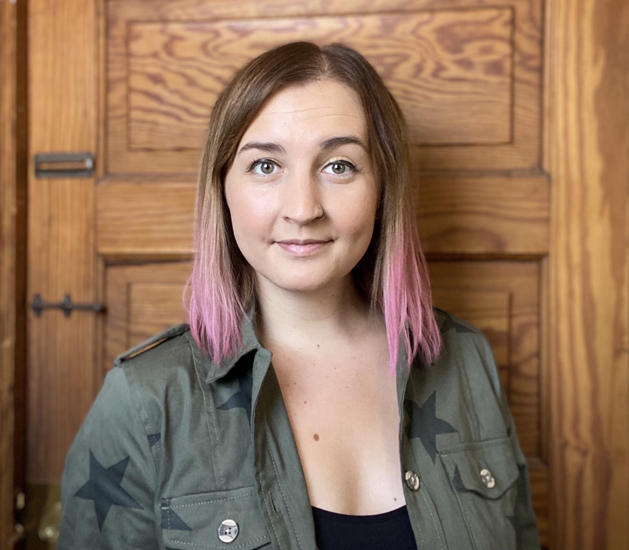 Headshot of Katie Kiesewetter. She is standing in front of of a wooden door with pink hair and an army green jacket with stars on it.