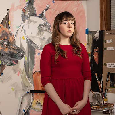 Caitlyn Doran stands before a painting of dogs in a red dress. Brunette hair falls on her shoulders and cropped bangs lay across her forehead.