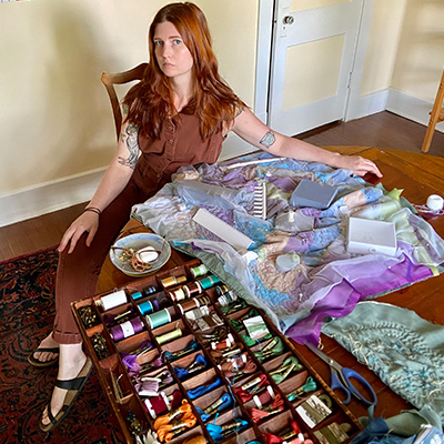 Millicent Kennedy sitting at a table with a box full of colorful threads and materials. Textile art in the making. Millicent wears a brown jumpsuit. Golden red hair, parted in the middle falls past her shoulders. 