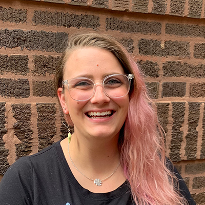 Taylor Whyte stands before a brick wall in her headshot. She wears a black T-shirt with a blue cat on it. Clear frame glasses sit on her nose and light pink hair is brushed to rest on her left shoulder.