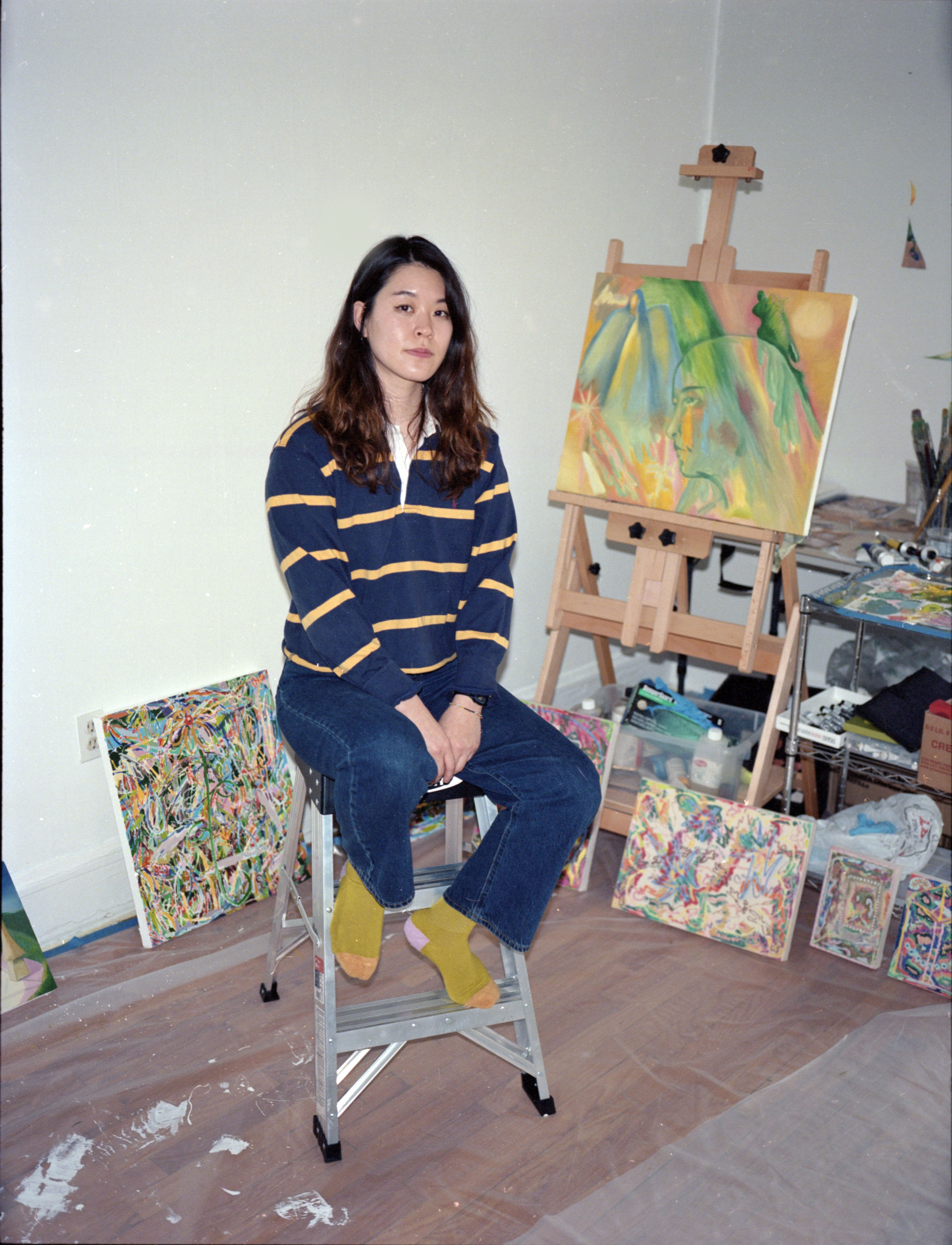 Michelle Chun sits on the top step of a three step ladder in front of a collection of in-progress paintings. She is wearing a striped shirt, jeans, and yellow socks.