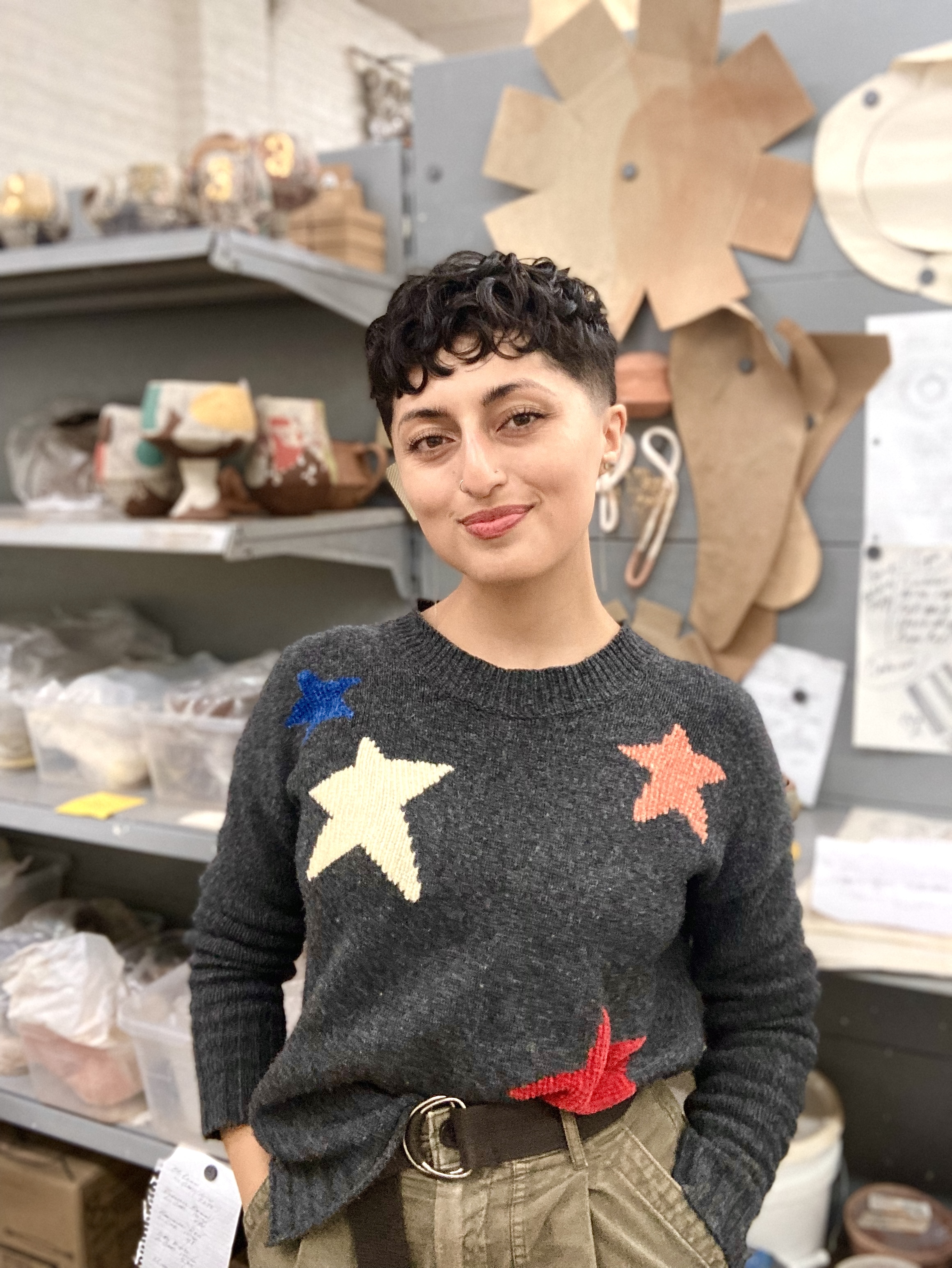 Headshot of Breana Marie Ferreira. The artist is wearing a gray sweater with large stars and is standing in front of a shelf of ceramic materials with her hands in her pockets.