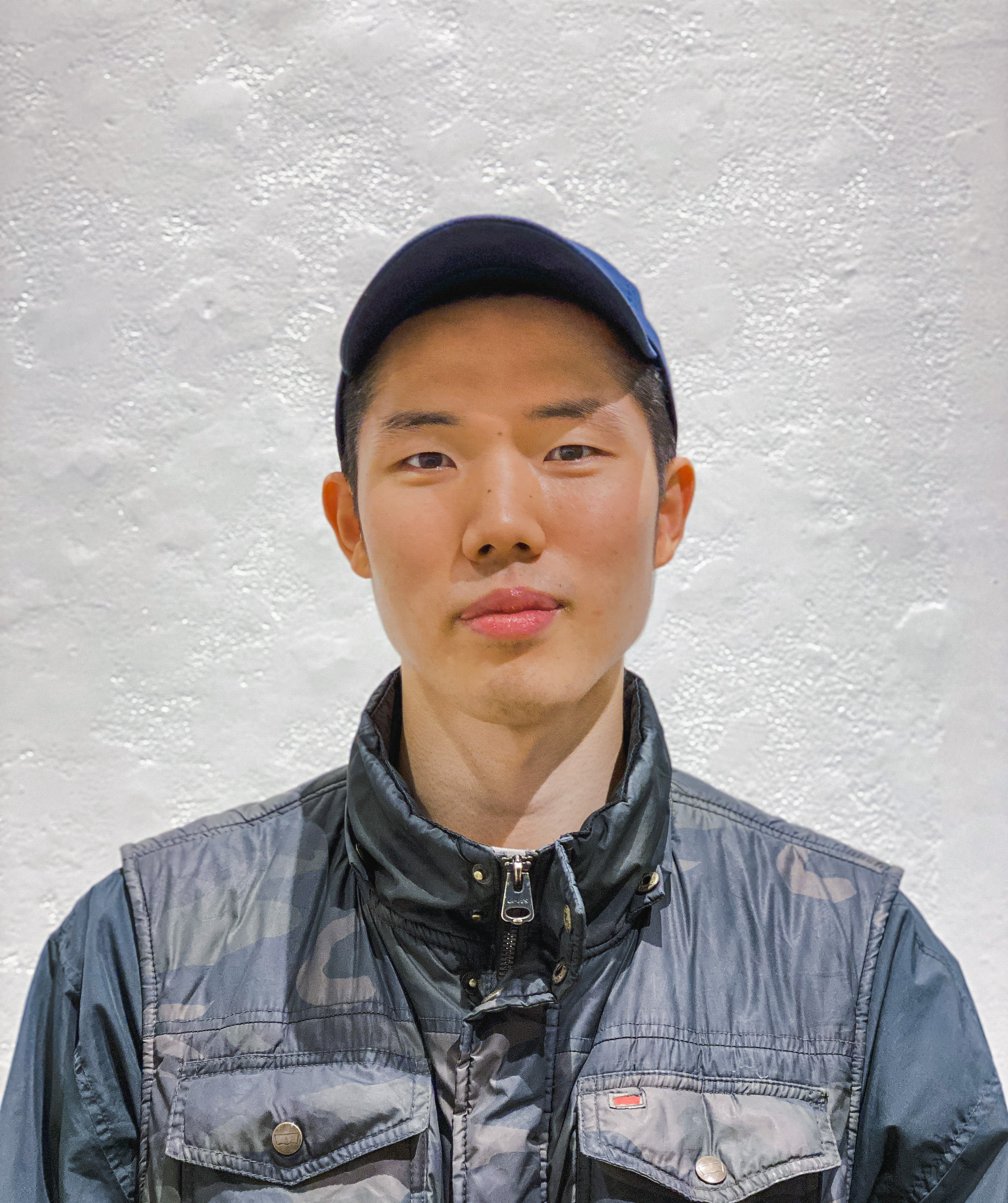 Headshot of Seuil Chung. He is wearing blue camouflage puffer vest, a blue baseball cap, and is standing in front of a textured white wall.
