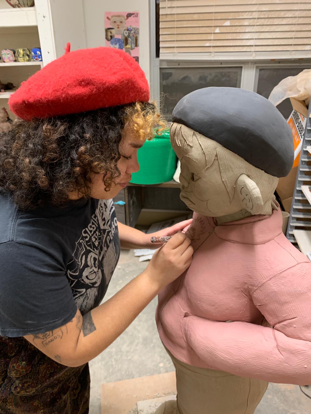 Sydnie Jimenez carving into a large-scale figure. The figure wears a pink jacket and a black hat. Jimenez is swearing a black t-shirt and a red beret.