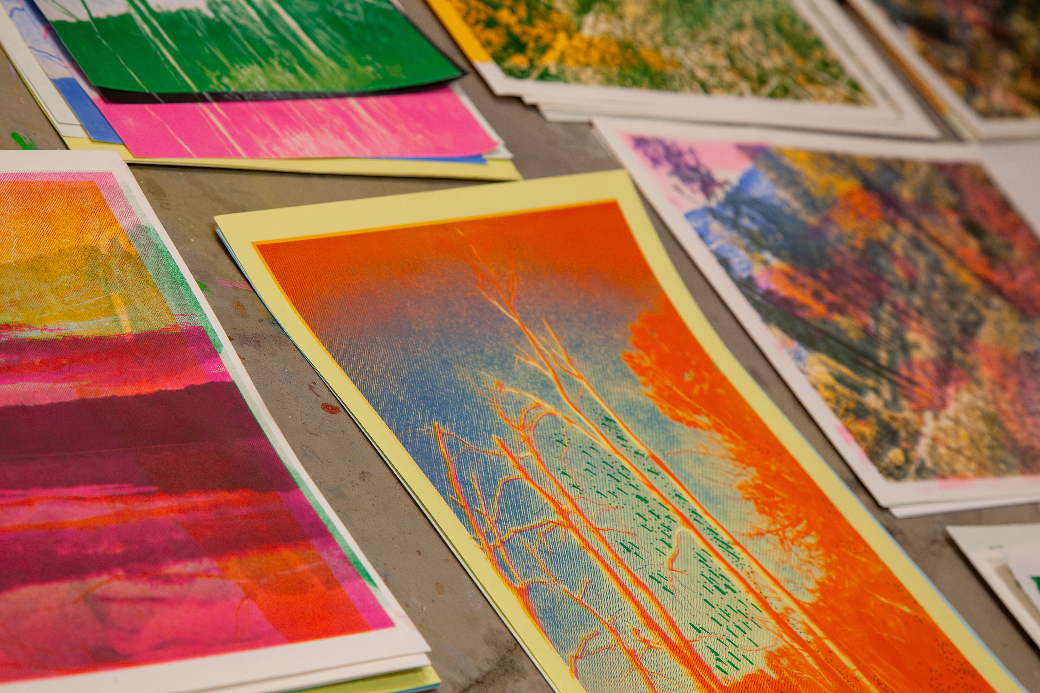 A selection of risograph prints in blue, orange, pink, and green. The main image in the center is of a group of leafless trees. 