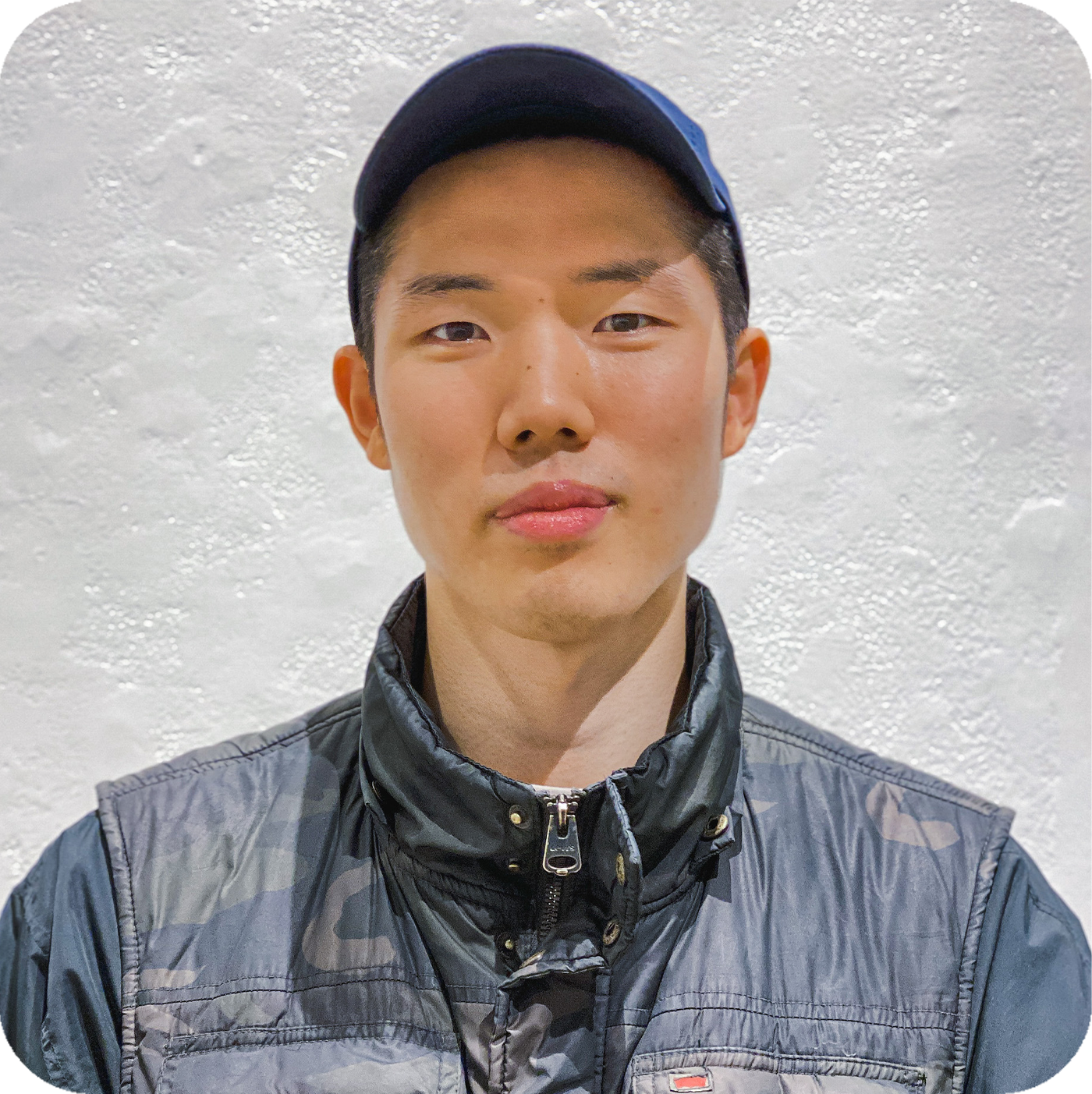 Headshot of Seuil Chung. He is wearing blue camouflage puffer vest, a blue baseball cap, and is standing in front of a textured white wall.